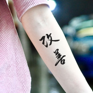 May include: A black temporary tattoo featuring two Chinese characters on a person's arm. The arm is pale, and the tattoo is in a bold, calligraphic style. The person is wearing a pink long-sleeved shirt.
