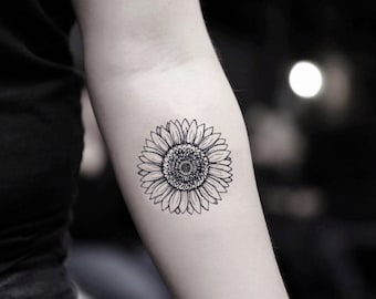 Puede incluir: Un diseño de tatuaje de girasol en blanco y negro en un brazo. El tatuaje presenta pétalos detallados que rodean un centro texturizado. La imagen es un primer plano, que destaca el intrincado trabajo de línea del diseño floral.