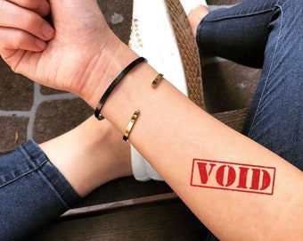 Può includere: Un braccio con un bracciale nero e un bracciale dorato. Un timbro rosso con la parola "VOID" è sul braccio. La persona indossa jeans blu e scarpe da ginnastica bianche.