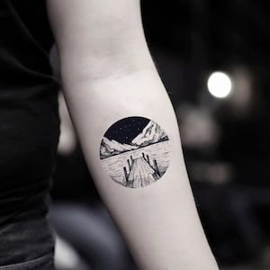 Pode incluir: Tatuagem a preto e branco de uma cena de paisagem dentro de um círculo. O desenho apresenta um cais de madeira que se estende para um lago, com montanhas ao fundo sob um céu estrelado.