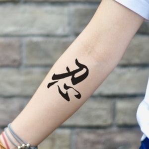 May include: Black kanji symbol temporary tattoo on the forearm. The tattoo is a bold, brushstroke style design. The arm is fair-skinned, and the background is a brick wall.