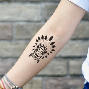 May include: A black temporary tattoo of a Native American chief with a feathered headdress. The tattoo is applied to the forearm, showcasing the detailed design of the face and headdress. The tattoo is a bold, graphic design.
