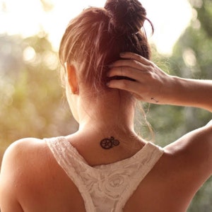 Könnte beinhalten: Ein schwarzes Zahnrad-Tattoo auf dem Nacken einer Person. Die Person trägt ein weißes Spitzentop. Das Haar der Person ist zu einem Dutt gebunden. Ein kleines Pfeiltattoo ist am Handgelenk der Person sichtbar.