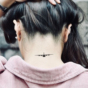Könnte beinhalten: Schwarzes Flugzeug-Tattoo am Nacken. Das Tattoo ist eine detaillierte Silhouette eines Flugzeugs mit Flügeln und mehreren Triebwerken. Die Person trägt ein rosa Cordhemd.