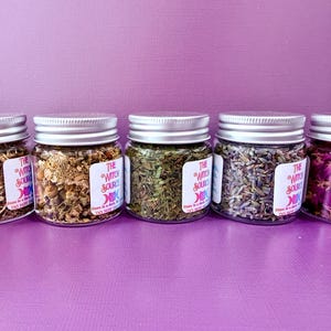 May include: Five small glass jars with silver lids, filled with dried herbs and flowers. The jars are labeled with white stickers that read "The Witch Source". The contents include lavender, rose petals, and other botanicals.