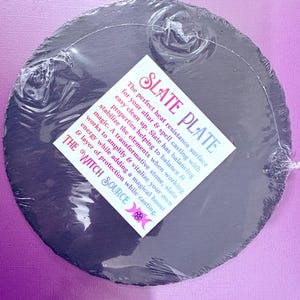 May include: A round, dark gray slate plate, packaged in clear plastic. The plate has a textured surface and a white label with the text "SLATE PLATE" and "THE WITCH SOURCE". The label describes the plate's use for altars and spell casting.