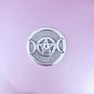 May include: A circular silver-toned metal medallion featuring a central five-pointed star enclosed within a circle, flanked by two crescent moons on the left and right sides. The background of the medallion has a textured, dotted surface finish.