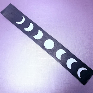 May include: A long, rectangular black wooden strip featuring a series of white painted moon phases. The design displays the lunar cycle, starting with a crescent moon, progressing through various waxing phases to a full moon, and ending with waning phases.