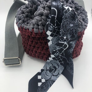 Puede incluir: Un bolso de cubo de crochet gris y burdeos con una bufanda de estampado floral blanco y negro. El bolso tiene una correa larga gris.