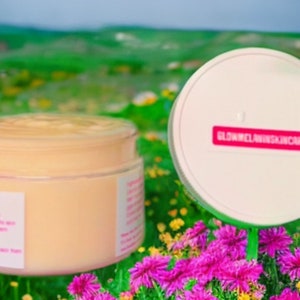 May include: A white jar with a pink label that says "GlowMelaninSkincare" filled with a light orange cream. The jar is sitting on a bed of pink and yellow flowers.