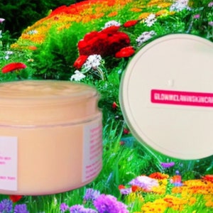 May include: A white jar with a pink label that says "GlowMelaninSkincare" filled with a light orange cream. The jar is sitting on a bed of colorful flowers.