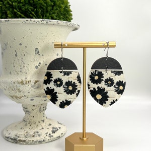 May include: A pair of black and white floral dangle earrings with cork backing. The earrings are displayed on a gold stand.