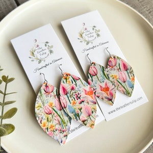 May include: A pair of floral print leather earrings with silver hooks. The earrings are shaped like a teardrop and feature a white background with pink, yellow, and blue flowers. The earrings are handcrafted in Arkansas and are available for purchase on the website www.theleathertulip.com.