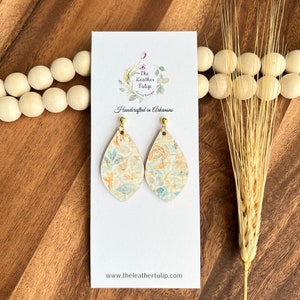 May include: Teardrop-shaped earrings with a floral pattern in shades of blue, beige, and orange. The earrings are displayed on a white card with the text "The Leather Tulip" and "Handcrafted in Arkansas". The earrings have gold-colored posts.