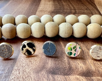 May include: Five pairs of round cork earrings with gold metal settings. The earrings feature different patterns, including a wood grain, leopard print, black and white, floral, and a speckled pattern.