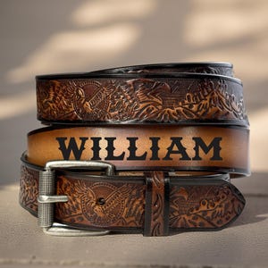 May include: Stacked leather belt with the name "WILLIAM" in bold black letters. The top and bottom belts feature an embossed eagle design. The buckle is silver-colored.