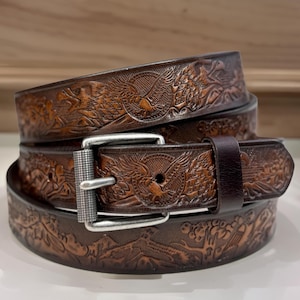 May include: A brown leather belt with an intricate eagle design embossed on the surface. The belt has a silver buckle and is layered with two additional belts.