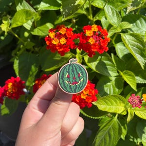 Könnte beinhalten: Ein runder Holzpin mit einem grünen Wassermelonen-Design und einem geschnitzten Kürbisgesicht. Der Pin befindet sich vor einem Hintergrund aus roten und gelben Blumen und grünen Blättern.