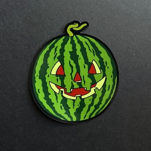 Puede incluir: Una pegatina redonda con un diseño de sandía verde tallada en forma de cara de calabaza. La calabaza tiene ojos y boca rojos, con un contorno amarillo. La pegatina tiene un tallo verde y está sobre un fondo negro.