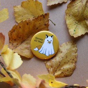May include: A yellow button with a cartoon fox wearing a white ghost costume. The button says "Spooky All Year" in black text.