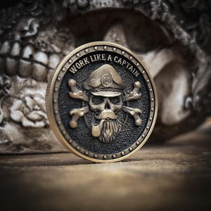 Puede incluir: Una moneda de color bronce con un diseño de calavera que lleva un gorro de capitán, huesos cruzados y una pipa. El borde de la moneda tiene pequeños remaches y el texto "WORK LIKE A CAPTAIN" está grabado. El fondo es una calavera borrosa.