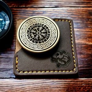 May include: A brass compass with a Celtic knot design and runes around the edge. The compass is on a brown leather wallet with a stitched border.