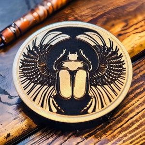 Puede incluir: Un posavasos redondo de metal dorado con un diseño negro y dorado de un escarabajo con alas. El escarabajo está centrado en el posavasos y tiene un diseño detallado.