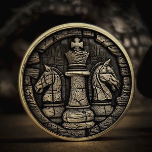 May include: A detailed, bronze-colored chess coin featuring a king piece flanked by two knight pieces. The coin has a textured, antique finish, with a dark background that highlights the chess pieces. The coin's edge is a lighter gold color.