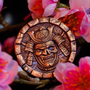 May include: A circular bronze-toned medallion featuring a detailed relief of a samurai mask with a skull-like face and traditional helmet. The medallion has a textured border and is surrounded by soft-focus pink cherry blossom flowers.