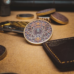 Puede incluir: Una moneda de metal dorado con un diseño detallado de una mariposa con patrones intrincados. La mariposa está en tonos de marrón, naranja y azul.