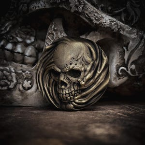 Puede incluir: Un medallón de calavera dorado, de estilo antiguo y detallado, con detalles de tela fluida. La calavera tiene una sonrisa detallada con la boca abierta. El fondo presenta una calavera tallada en piedra y una superficie oscura y texturizada.