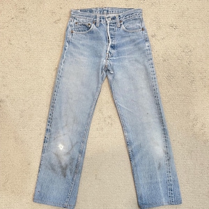 May include: Light blue denim jeans with a button fly, five pockets, and a faded appearance. The jeans show wear and tear, including a small hole on the left leg. The jeans are laid flat on a neutral surface.