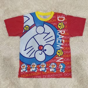 May include: Red and blue Doraemon t-shirt with a large Doraemon graphic on the front. The shirt has a yellow collar and red sleeves. The word "Doraemon" is printed vertically on the right side of the shirt.