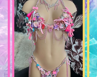 Puede incluir: Un conjunto de bikini rosa y plateado con adornos temáticos de Barbie. La parte superior e inferior están adornadas con dijes, lazos y muñecas Barbie en miniatura. El collar deletrea "Barbie" en letras plateadas.