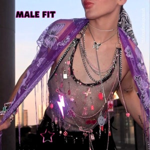 Peut inclure: Une personne portant un haut en maille noire avec une variété de chaînes, de breloques et de pendentifs argentés et roses. Le haut est superposé à une écharpe violette avec des broderies florales. La personne porte également un pantalon noir et un accessoire en forme d'étoile.