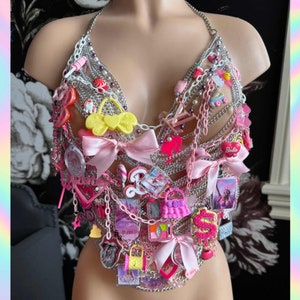 Pode incluir: Um top de malha de corrente prateada adornado com laços rosa, amuletos e acessórios em miniatura. Os amuletos incluem mini bolsas, doces e o logotipo da Barbie. O top é estilo halter.