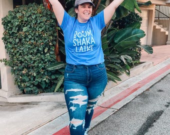 May include: A person wearing a blue t-shirt with the text "BOOM SHAKA LAIKA" in white. They are wearing ripped blue jeans and white trainers. The person is standing on a pavement with a red line.