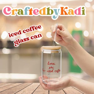 May include: Clear glass can with a bamboo lid and a clear straw, designed for iced coffee. The can has red text that reads "borei pri ha-iced coffee". The top of the image has the text "CraftedbyKadi" in colorful letters. The words "iced coffee glass can" are also in red text.