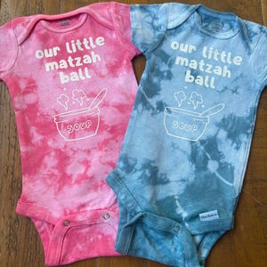 May include: Two tie-dye baby onesies, one pink and one blue, with the text "our little matzah ball" and a cartoon bowl of soup.