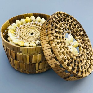 Può includere: Un cesto rotondo intrecciato con un coperchio, entrambi realizzati in materiale intrecciato marrone naturale. Il cesto è riempito con una disposizione circolare di perline di conchiglia bianche e gialle.