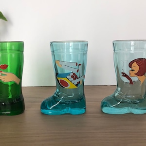Op de afbeelding: Drie glazen shotglazen in de vorm van laarzen. De glazen zijn versierd met verschillende afbeeldingen. Een glas is groen en heeft een hand die een sigaret vasthoudt. Een ander glas is blauw en heeft een vrouw die een sigaret rookt. Het derde glas is blauw en heeft een doosje sigaretten.