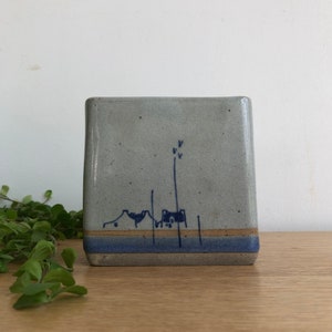 Può includere: Un piccolo vaso quadrato in ceramica grigia con un disegno dipinto a mano in blu e bianco di case e un albero. Il vaso ha uno sfondo grigio chiaro con una sottile striscia blu vicino al fondo.