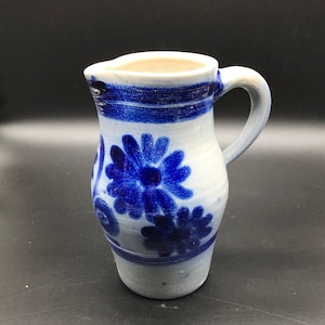 Op de afbeelding: Een witte keramische kan met een blauw bloemenmotief. De kan heeft een rond lichaam en een handvat.