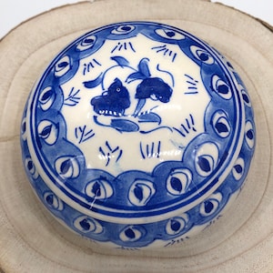 May include: A small, round, blue and white ceramic trinket box with a lid. The lid features a design of two stylised peacocks with their tails spread out. The box is decorated with a blue and white peacock feather pattern.