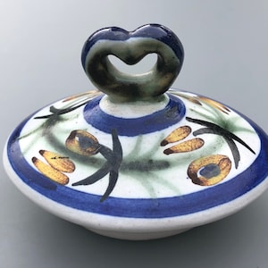 Può includere: Un coperchio in ceramica bianca con un motivo floreale blu e verde e un manico a forma di cuore. Il manico è smaltato in blu scuro e marrone.