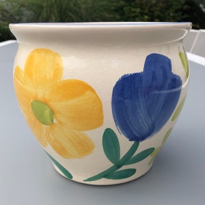 Può includere: Un vaso di ceramica bianca con un bordo blu e un disegno floreale dipinto. I fiori sono gialli, blu e verdi.