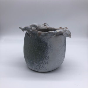 May include: A white ceramic vase with a textured, uneven surface and a green glaze. The vase has a unique, organic shape with a wide opening and a rounded bottom.