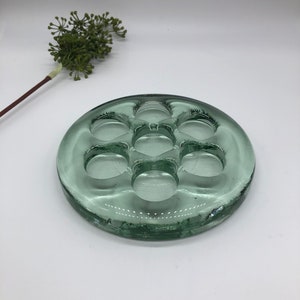 May include: A round, clear green glass flower frog with seven holes for arranging flowers.