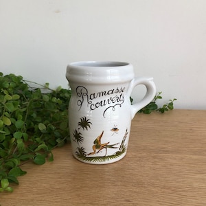 Könnte beinhalten: Weißer Keramik Krug mit Henkel, mit einer grünen und braunen Illustration eines Vogels und Pflanzen. Der Krug trägt den Text "Ramasse couverts" in schwarzer Kursivschrift.