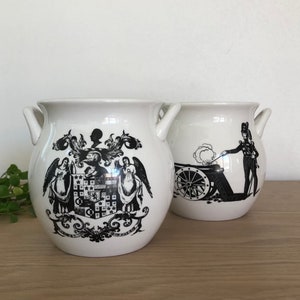 Op de afbeelding: Twee witte keramische potten met zwart-witte ontwerpen. De ene pot heeft een wapenschild met engelen en een schild. De andere pot heeft een soldaat die een kanon afvuurt.